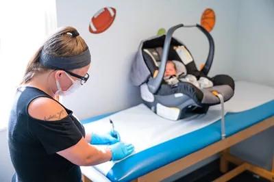 a medical provider writing in a chart with a baby in a baby carrier nearby